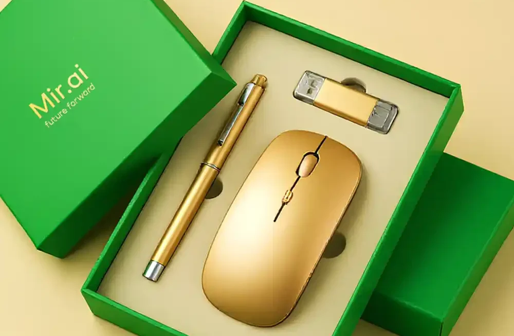 A high-angle studio shot displays a sleek corporate gift set arranged in a vibrant green box against a smooth, pale beige background. The box is open, revealing a cream-colored foam insert with custom cutouts holding three matching gold-colored office accessories: a slender ballpoint pen on the left, a minimalist wireless mouse in the center, and a rectangular USB flash drive with clear caps positioned at the top right. The box's lid, resting to the left of the base, features the branding "Mir.ai" followed by the slogan "future forward" printed in elegant gold lettering. The lighting is soft and even, highlighting the metallic sheen of the gold items and the matte texture of the green packaging. Corporate Gifting Boxes