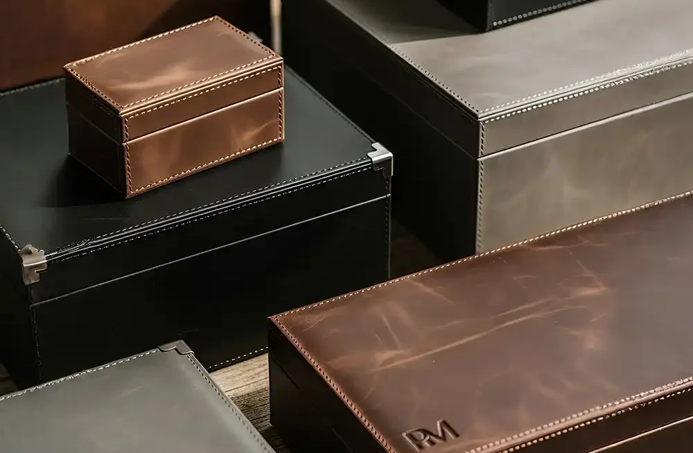 A high-angle, studio-style shot displays a variety of stacked and scattered leather storage boxes made by Creative Space, resting on a weathered wooden table. The organizers appear in three distinct colors—rich cognac brown, matte black, and slate grey—each showcasing a premium finish with visible grain and distinct stitching along the seams. Several of the larger boxes feature reinforced silver metal corners, adding a structured, vintage aesthetic. The lighting is soft and directional, casting gentle shadows that emphasize the tactile quality of the leather and the embossed detailing on the front-most brown box, presenting a refined solution for workspace organization. Premium Leather Boxes