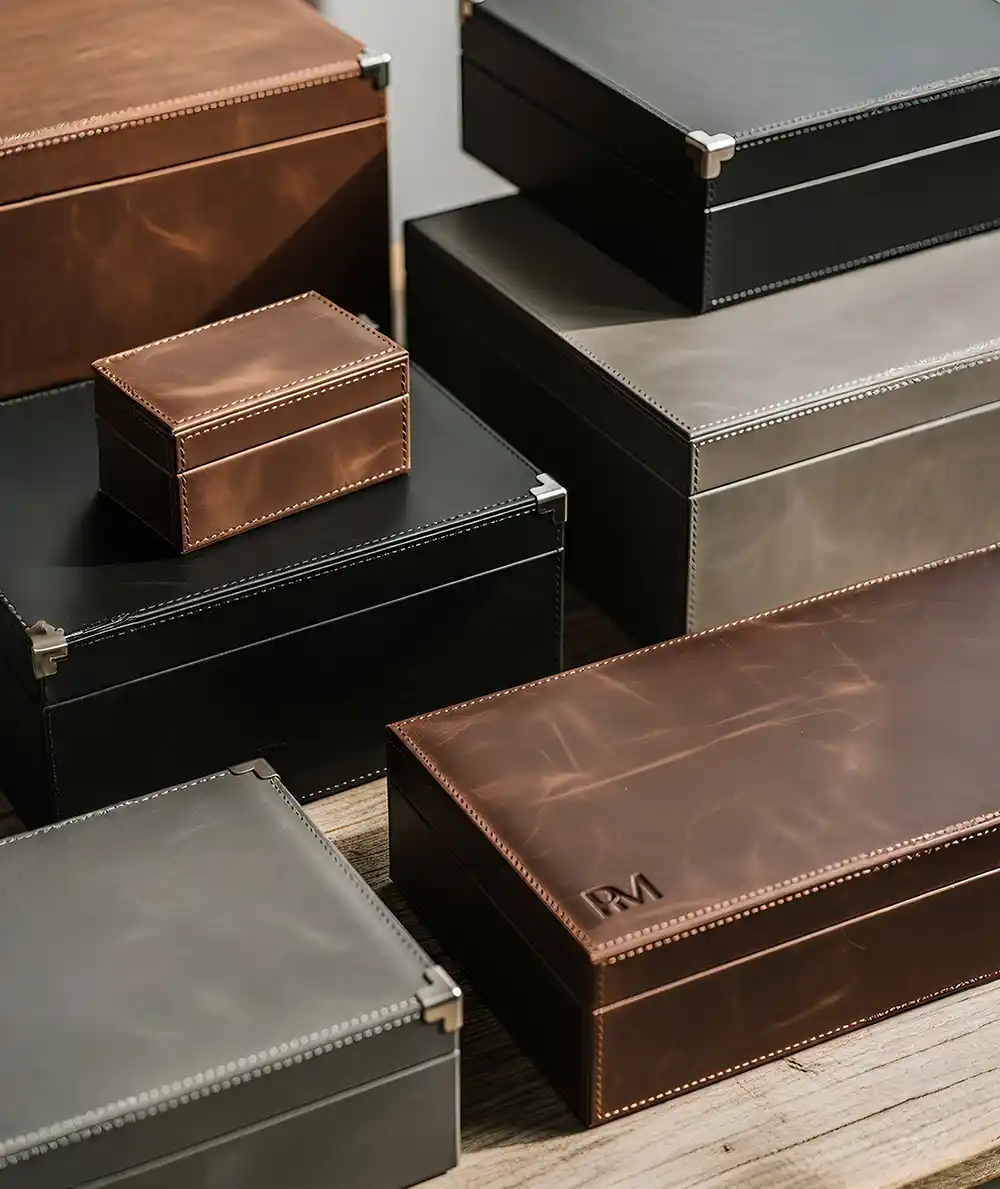 A high-angle, studio-style shot displays a variety of stacked and scattered leather storage boxes made by Creative Space, resting on a weathered wooden table. The organizers appear in three distinct colors—rich cognac brown, matte black, and slate grey—each showcasing a premium finish with visible grain and distinct stitching along the seams. Several of the larger boxes feature reinforced silver metal corners, adding a structured, vintage aesthetic. The lighting is soft and directional, casting gentle shadows that emphasize the tactile quality of the leather and the embossed detailing on the front-most brown box, presenting a refined solution for workspace organization. Premium Leather Boxes