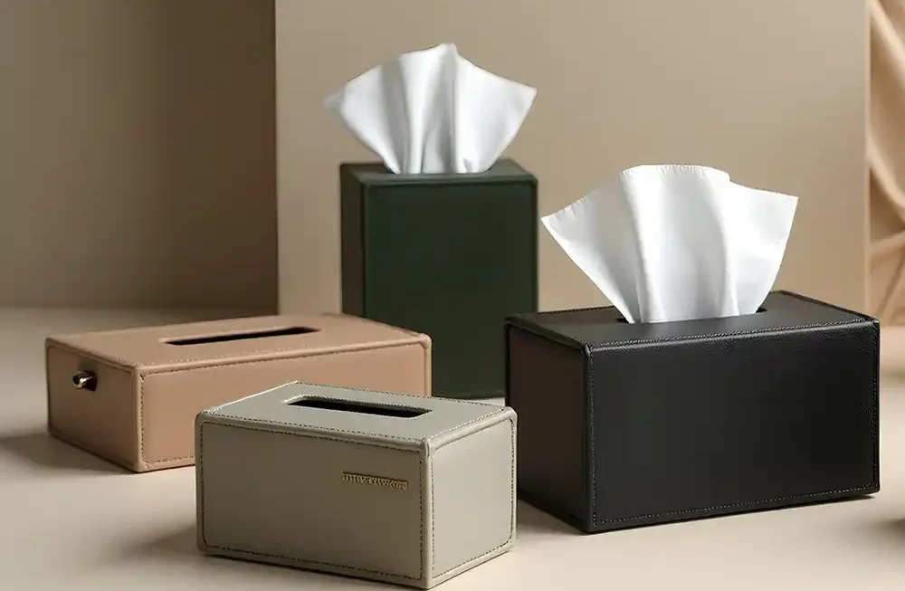 A polished, high-angle studio shot showcases a collection of premium leather tissue box covers made by Creative Space, arranged on a smooth beige surface against a minimalist architectural background. The assortment includes four distinct pieces: a tall, dark green cube and a rectangular black box, both featuring tufts of white tissue emerging from the top, alongside a warm tan rectangular box and a smaller light grey holder in the foreground. The empty grey and tan boxes reveal clean oval openings, while the grey box is accented with a small gold metal logo plate, highlighting the refined stitching and sleek design of the home accessories. A high-angle studio photograph displays a luxurious collection of velvet jewelry and gift boxes made by Creative Space, artistically stacked and arranged on a warm wooden table. The boxes come in a variety of sizes and deep jewel tones, including emerald green, royal blue, vibrant red, and slate grey. Several boxes are open, revealing sophisticated interiors that feature either fitted velvet inserts or gathered satin linings in contrasting blue shades, accented by small gold push-button clasps and gold foil branding inside the lids. The soft lighting emphasizes the plush texture of the exterior fabric and the elegant gold trim detailing on the larger, closed containers at the base. Décor Tissue Holders
