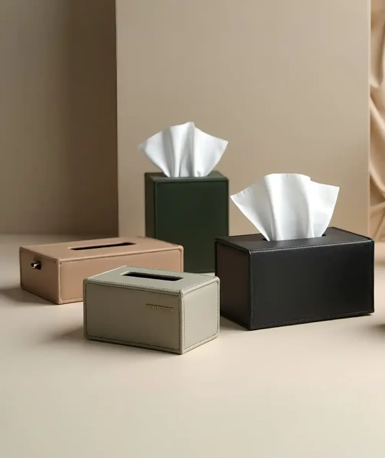 A polished, high-angle studio shot showcases a collection of premium leather tissue box covers made by Creative Space, arranged on a smooth beige surface against a minimalist architectural background. The assortment includes four distinct pieces: a tall, dark green cube and a rectangular black box, both featuring tufts of white tissue emerging from the top, alongside a warm tan rectangular box and a smaller light grey holder in the foreground. The empty grey and tan boxes reveal clean oval openings, while the grey box is accented with a small gold metal logo plate, highlighting the refined stitching and sleek design of the home accessories. A high-angle studio photograph displays a luxurious collection of velvet jewelry and gift boxes made by Creative Space, artistically stacked and arranged on a warm wooden table. The boxes come in a variety of sizes and deep jewel tones, including emerald green, royal blue, vibrant red, and slate grey. Several boxes are open, revealing sophisticated interiors that feature either fitted velvet inserts or gathered satin linings in contrasting blue shades, accented by small gold push-button clasps and gold foil branding inside the lids. The soft lighting emphasizes the plush texture of the exterior fabric and the elegant gold trim detailing on the larger, closed containers at the base. Décor Tissue Holders
