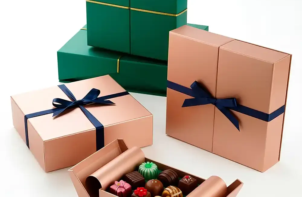 A sophisticated studio shot displays an arrangement of high-end gift packaging for chocolates or small luxury items on a clean white surface. The foreground features an open rose gold box tied with a dark navy satin ribbon, with the sides unfolded to dramatically present a tray of colorful, artisan bonbons and truffles. Surrounding it are several closed boxes: a tall vertical rose gold box and a flat square rose gold box, both accented with the navy ribbon, and a stack of deep emerald green boxes sealed with a delicate gold metallic string in the background, conveying a high degree of luxury and elegance suitable for celebratory gifting. Miniature Gift Boxes