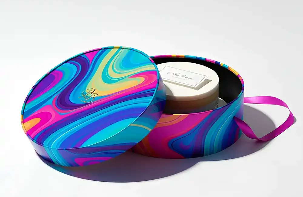 An overhead studio shot shows an open round gift box with a vibrant, colorful, psychedelic, swirled pattern lid and a white box with a gold foiled "Bb" logo in a cursive font placed inside it. The box has a black interior and a dark pink ribbon coming out of it. The gift box is sitting on a seamless white surface, creating a clean, bright, and professional aesthetic. Pastry & Bakery Boxes