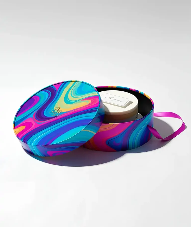 An overhead studio shot shows an open round gift box with a vibrant, colorful, psychedelic, swirled pattern lid and a white box with a gold foiled "Bb" logo in a cursive font placed inside it. The box has a black interior and a dark pink ribbon coming out of it. The gift box is sitting on a seamless white surface, creating a clean, bright, and professional aesthetic. Pastry & Bakery Boxes