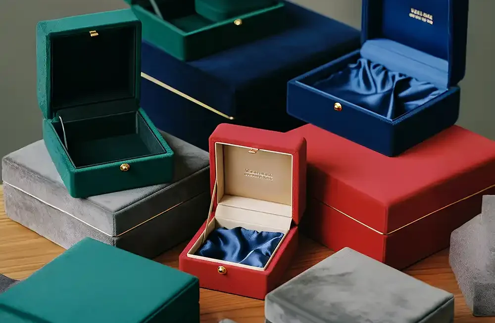 A high-angle studio photograph displays a luxurious collection of velvet jewelry and gift boxes made by Creative Space, artistically stacked and arranged on a warm wooden table. The boxes come in a variety of sizes and deep jewel tones, including emerald green, royal blue, vibrant red, and slate grey. Several boxes are open, revealing sophisticated interiors that feature either fitted velvet inserts or gathered satin linings in contrasting blue shades, accented by small gold push-button clasps and gold foil branding inside the lids. The soft lighting emphasizes the plush texture of the exterior fabric and the elegant gold trim detailing on the larger, closed containers at the base. Velvet Finish Boxes