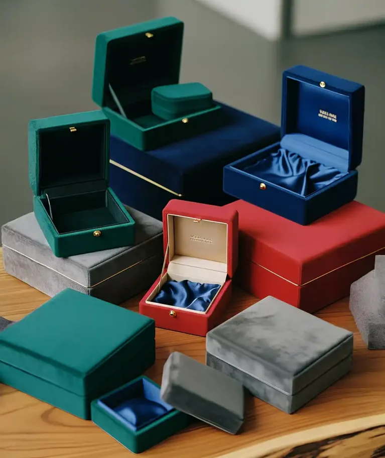 A high-angle studio photograph displays a luxurious collection of velvet jewelry and gift boxes made by Creative Space, artistically stacked and arranged on a warm wooden table. The boxes come in a variety of sizes and deep jewel tones, including emerald green, royal blue, vibrant red, and slate grey. Several boxes are open, revealing sophisticated interiors that feature either fitted velvet inserts or gathered satin linings in contrasting blue shades, accented by small gold push-button clasps and gold foil branding inside the lids. The soft lighting emphasizes the plush texture of the exterior fabric and the elegant gold trim detailing on the larger, closed containers at the base. Velvet Finish Boxes