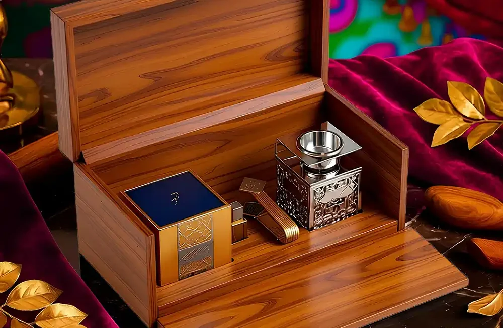 A highly dramatic, opulent studio shot showcases a premium wooden gift set, likely for incense or fragrance, meticulously crafted and made by Creative Space. The focal point is the large box made of polished, reddish-brown wood, displayed open with the front panel lowered to serve as a presentation tray. Inside the wooden compartments sits a decorative metal bakhoor burner with intricate filigree, positioned next to a dark blue square container (presumably for the oud or incense) and smaller gold accessories. The entire scene is styled lavishly on a dark marble surface draped with rich magenta velvet, accented by scattered gold leaves and a vivid, colorful curtain backdrop, enhancing the luxurious, Eastern-inspired aesthetic. Wooden Presentation Boxes