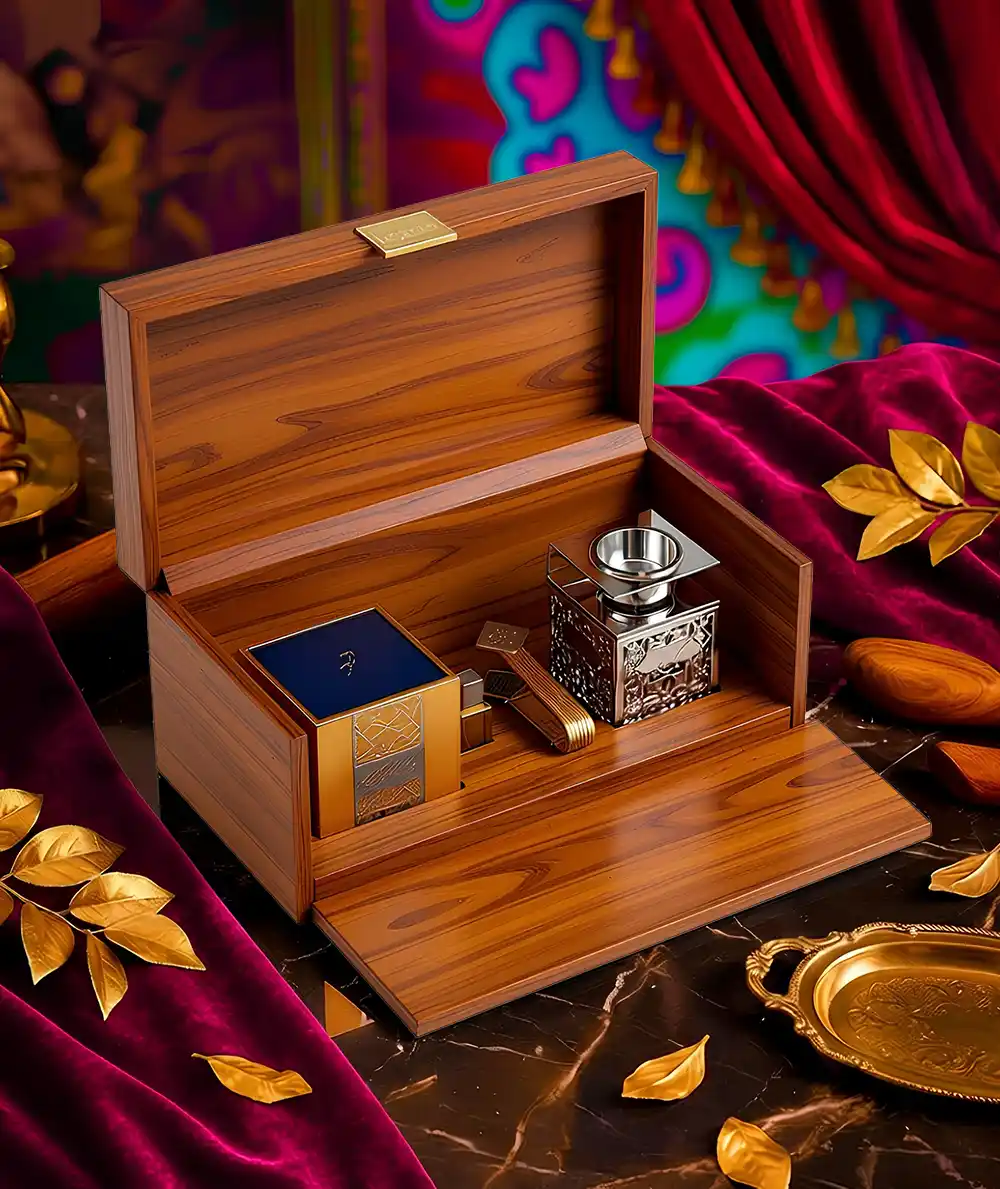 A highly dramatic, opulent studio shot showcases a premium wooden gift set, likely for incense or fragrance, meticulously crafted and made by Creative Space. The focal point is the large box made of polished, reddish-brown wood, displayed open with the front panel lowered to serve as a presentation tray. Inside the wooden compartments sits a decorative metal bakhoor burner with intricate filigree, positioned next to a dark blue square container (presumably for the oud or incense) and smaller gold accessories. The entire scene is styled lavishly on a dark marble surface draped with rich magenta velvet, accented by scattered gold leaves and a vivid, colorful curtain backdrop, enhancing the luxurious, Eastern-inspired aesthetic. Wooden Presentation Boxes