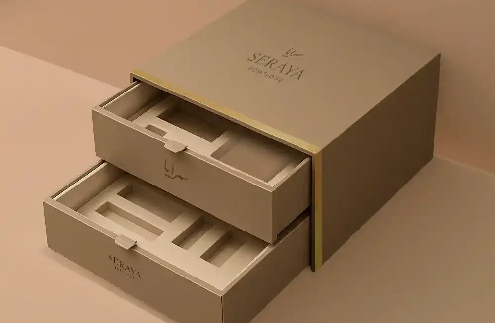 A minimalist, high-angle studio photograph features a sophisticated, two-tier custom gift box made by Creative Space, constructed from a matte, taupe-colored rigid paperboard. The design is presented with both horizontal sliding drawers partially pulled out, revealing custom-fitted inserts with multiple rectangular cutouts for product presentation. The top surface of the main body is prominently branded with the logo "SERAYA BOUTIQUE" and accented by a thin gold metallic trim that wraps around the corner. The box rests on a clean, monochromatic surface, where the lighting uses subtle shadows to emphasize the modern, clean-lined structure of the empty luxury packaging. A highly dramatic, opulent studio shot showcases a premium wooden gift set, likely for incense or fragrance, meticulously crafted and made by Creative Space. The focal point is the large box made of polished, reddish-brown wood, displayed open with the front panel lowered to serve as a presentation tray. Inside the wooden compartments sits a decorative metal bakhoor burner with intricate filigree, positioned next to a dark blue square container (presumably for the oud or incense) and smaller gold accessories. The entire scene is styled lavishly on a dark marble surface draped with rich magenta velvet, accented by scattered gold leaves and a vivid, colorful curtain backdrop, enhancing the luxurious, Eastern-inspired aesthetic. Customized Gift Boxes
