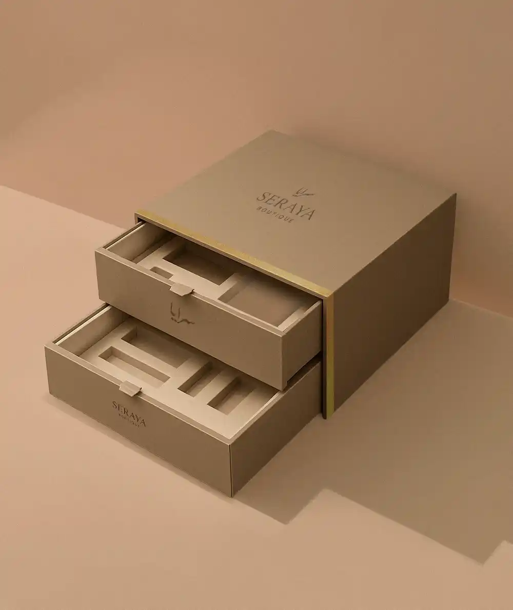 A minimalist, high-angle studio photograph features a sophisticated, two-tier custom gift box made by Creative Space, constructed from a matte, taupe-colored rigid paperboard. The design is presented with both horizontal sliding drawers partially pulled out, revealing custom-fitted inserts with multiple rectangular cutouts for product presentation. The top surface of the main body is prominently branded with the logo "SERAYA BOUTIQUE" and accented by a thin gold metallic trim that wraps around the corner. The box rests on a clean, monochromatic surface, where the lighting uses subtle shadows to emphasize the modern, clean-lined structure of the empty luxury packaging. A highly dramatic, opulent studio shot showcases a premium wooden gift set, likely for incense or fragrance, meticulously crafted and made by Creative Space. The focal point is the large box made of polished, reddish-brown wood, displayed open with the front panel lowered to serve as a presentation tray. Inside the wooden compartments sits a decorative metal bakhoor burner with intricate filigree, positioned next to a dark blue square container (presumably for the oud or incense) and smaller gold accessories. The entire scene is styled lavishly on a dark marble surface draped with rich magenta velvet, accented by scattered gold leaves and a vivid, colorful curtain backdrop, enhancing the luxurious, Eastern-inspired aesthetic. Customized Gift Boxes