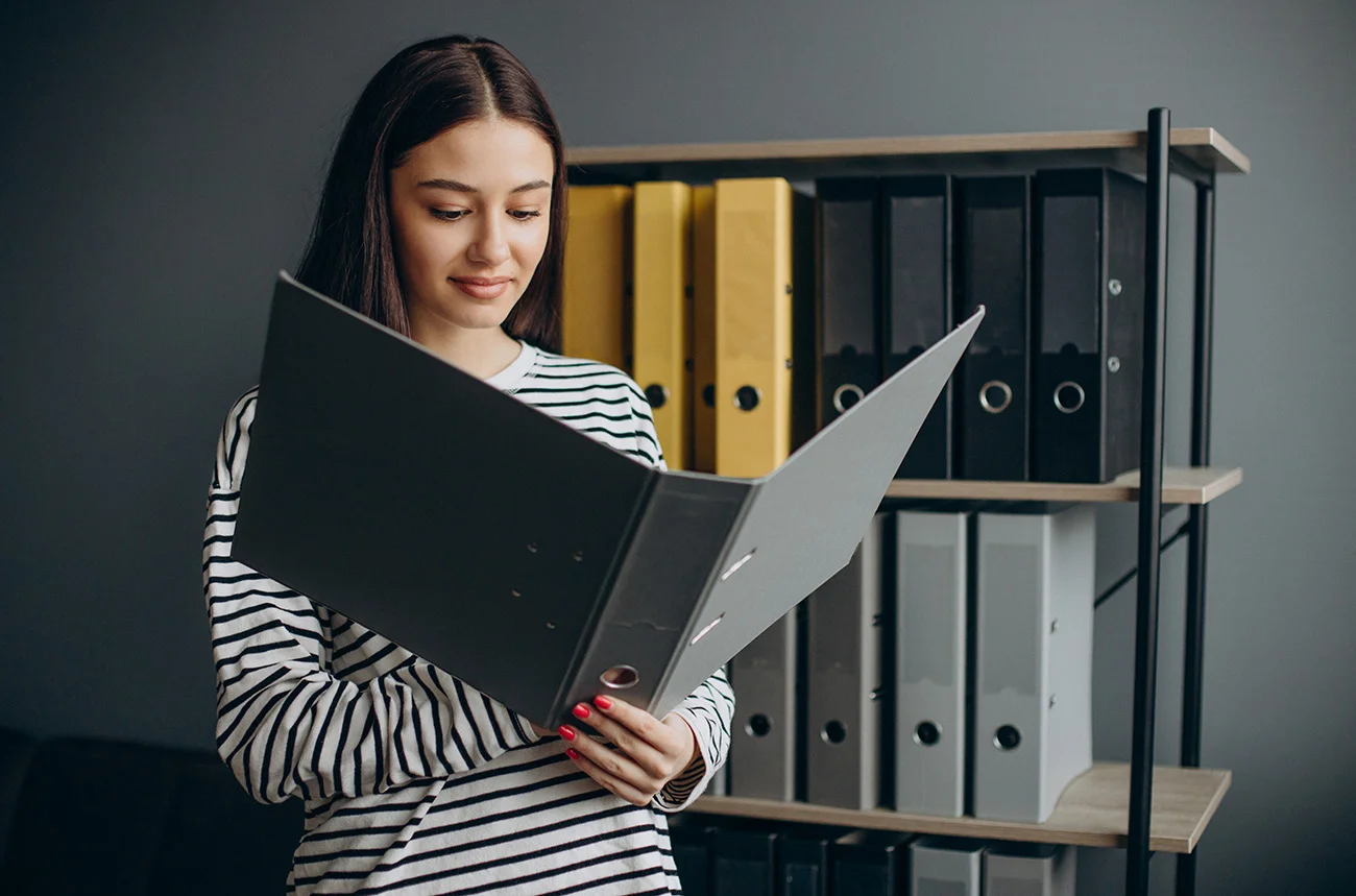 In this image, a young professional woman is seen in a modern workspace, reviewing documents within a large Creative Space Gifts organizational binder.Key Visual ElementsCorporate Organization: The woman, wearing a striped shirt, stands in front of a sleek black shelving unit filled with neatly arranged lever-arch binders in professional tones of mustard yellow, charcoal, and light gray.Functional Design: She holds a high-capacity, dark gray binder open, showcasing its durable construction—ideal for managing extensive project files or brand guidelines.Atmosphere: The minimalist gray background and clean lines emphasize a productive, organized "creative space" environment.Product Spotlight: Organizational ToolsFeatureDescriptionMaterialHeavy-duty rigid board with a smooth, matte finish.CapacityDesigned for high-volume document storage with metal ring mechanisms.AestheticsProfessional color-blocking that fits modern office decor.This setting highlights the transition from luxury gift presentation to practical office efficiency, providing a complete ecosystem for brand identity and physical organization.