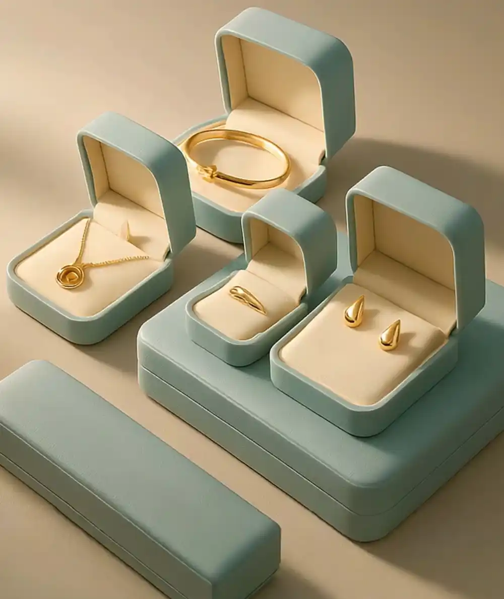 A high-angle, studio-quality shot displaying a cohesive set of luxury custom jewelry boxes packaging, made by Creative Space. The collection features a soft, matte sage-green exterior finish with plush, cream-colored interior lining. Four open boxes are arranged on a matching tiered platform, showcasing gold jewelry pieces: a sleek bangle bracelet in a square box, a circular pendant necklace in a rectangular box, a minimalist ring in a small square box, and gold teardrop earrings in a medium box. A long, closed rectangular box rests in the foreground on the left. The lighting is soft and warm, highlighting the smooth texture of the boxes and the gleam of the gold jewelry. Custom jewelry boxes UAE.