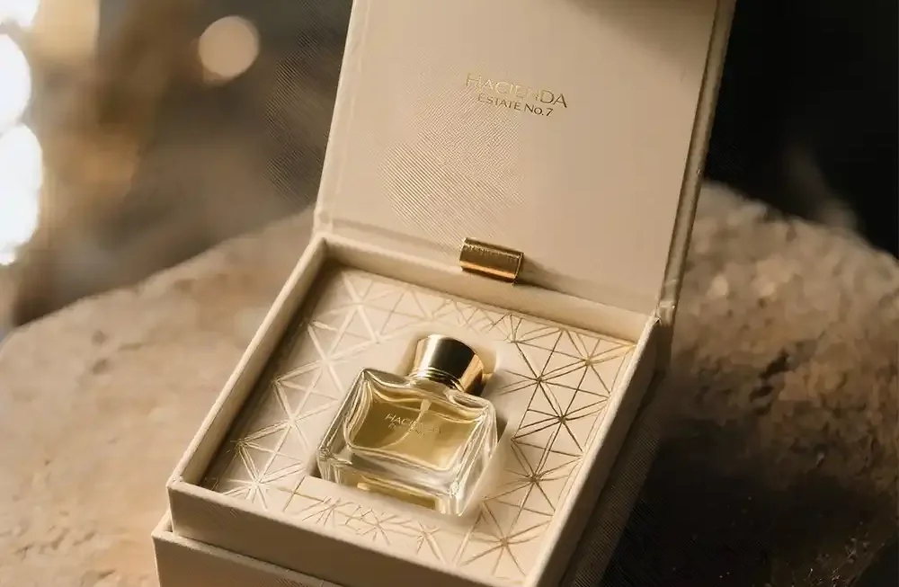 An exquisitely packaged perfume bottle nestled within an open, cream-colored luxury box. The box, made of a rigid material with a soft, leather-like texture, has a hinged lid that stands upright, revealing the perfume's name "HACIENDA ESTAE N°7" subtly embossed in gold on the inside. The perfume bottle itself is small, rectangular, and made of clear glass, filled with a warm, amber-toned liquid. It features a simple gold cap. The bottle rests on a cream-colored insert with a delicate, geometric gold pattern, adding to its sophisticated presentation. The entire box is positioned on a rustic, textured surface that appears to be rough-hewn wood or stone, with a soft, natural-toned fabric draped underneath, contrasting beautifully with the refined elegance of the perfume packaging. The lighting is soft and warm, highlighting the golden accents and the rich color of the perfume. Created by creative space. Premium perfume boxes in UAE