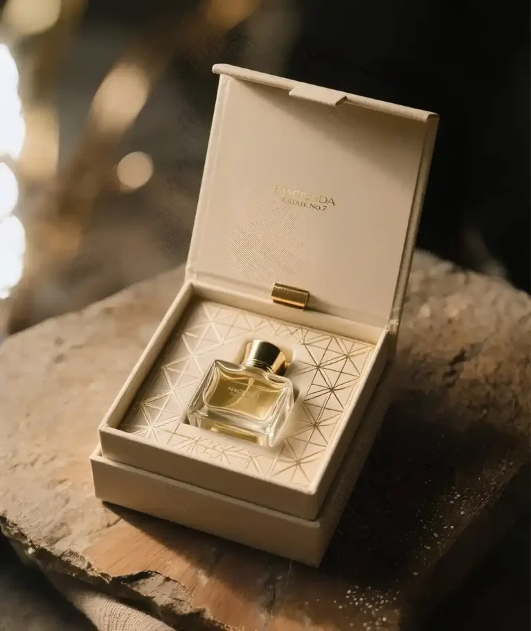 An exquisitely packaged perfume bottle nestled within an open, cream-colored luxury box. The box, made of a rigid material with a soft, leather-like texture, has a hinged lid that stands upright, revealing the perfume's name "HACIENDA ESTAE N°7" subtly embossed in gold on the inside. The perfume bottle itself is small, rectangular, and made of clear glass, filled with a warm, amber-toned liquid. It features a simple gold cap. The bottle rests on a cream-colored insert with a delicate, geometric gold pattern, adding to its sophisticated presentation. The entire box is positioned on a rustic, textured surface that appears to be rough-hewn wood or stone, with a soft, natural-toned fabric draped underneath, contrasting beautifully with the refined elegance of the perfume packaging. The lighting is soft and warm, highlighting the golden accents and the rich color of the perfume. Created by creative space. Premium perfume boxes in UAE
