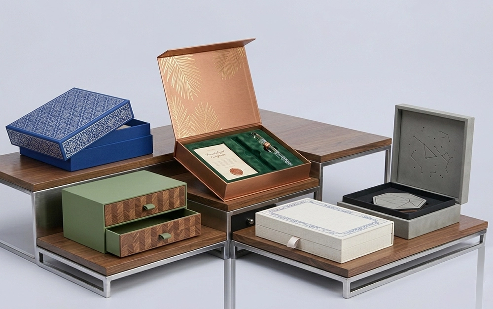 A medium shot shows six distinct gift boxes displayed on three tiered walnut and brushed metal tables against a plain white background. The top shelf on the left has a blue, patterned gift box that is slightly askew. The box on the second shelf has an open cover, with a brown color on the outside and gold details of palm tree leaves on the inside. It has a green interior with a pen and an invitation with the word "Invitation" written on top. The lower box on the left is a small green dresser with two wooden drawers that are open. There is a small, white cloth rectangular gift box on the table. On the top table to the right, there is a large, grey square-shaped open gift box with gold star formations inside the cover. It has a black interior with a grey pentagram shape on a coaster with gold details.