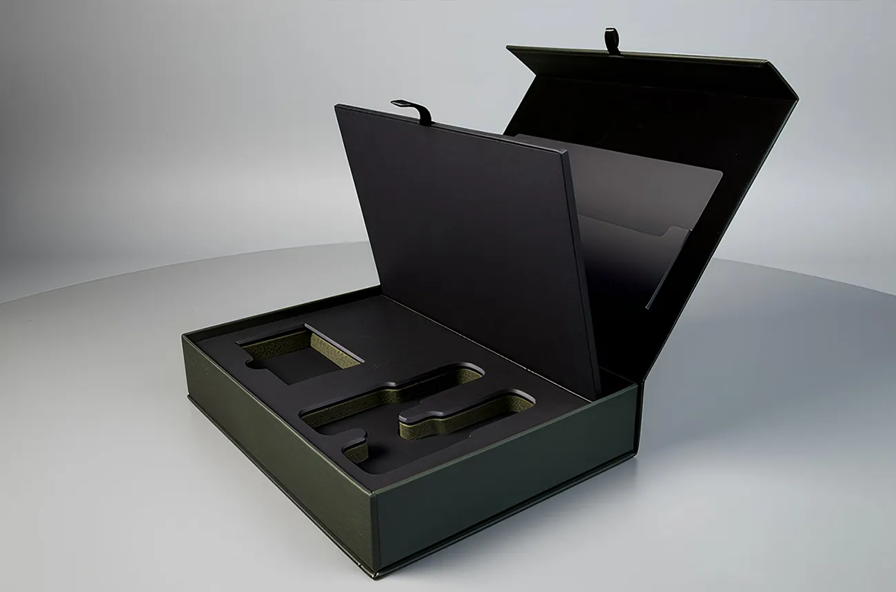 A detailed close-up shot of a luxury matte forest green rigid gift box from Creative Space Gifts, shown partially open to reveal a complex, multi-layered interior. The box features several high-end structural elements: Custom Die-Cut Foam Insert: A dark charcoal foam base with precise cutouts designed to securely hold specific luxury items. Integrated Document Holder: An interior flap with a small ribbon pull tab that lifts to reveal a dedicated compartment for brochures, certificates, or personal notes. Magnetic Flip-Top Lid: The lid is shown angled open, highlighting the sturdy construction and seamless hinge. Textured Exterior: The deep green paper finish has a subtle, premium texture that contrasts with the smooth, black interior lining. This packaging solution demonstrates a sophisticated "unboxing" experience, prioritizing both item protection and an elegant presentation for corporate or high-end retail gifts.
