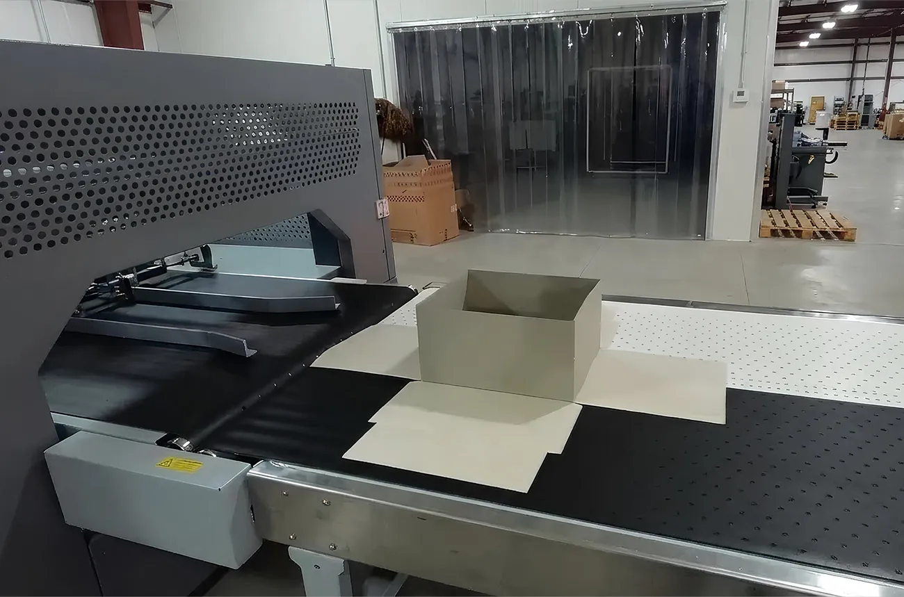 This image provides a behind-the-scenes look at the precision manufacturing process for Creative Space Gifts premium packaging. It captures a high-end rigid box at a critical stage of assembly on an automated production line.Manufacturing & Technical DetailsAutomated Assembly: A light beige rigid box sits on a wide, industrial conveyor belt. The machinery features a black non-slip surface and a perforated white transition plate, indicating a high-tech facility designed for precision folding and gluing.Structural Form: The image shows the box in a "semi-erected" state, where the main walls are formed while the exterior wrapping material (the "cover") is being precisely applied or folded.Industrial Setting: The background reveals a clean, spacious factory floor with additional machinery, heavy-duty shelving, and safety features like industrial plastic strip curtains.Quality Control: The sharp, clean angles of the box on the belt highlight the structural integrity and consistent quality that Creative Space Gifts maintains during large-scale production runs.Production SignificanceAspectDetailDurabilityHigh-thickness chipboard used for long-lasting rigid construction.ScaleFully automated line capable of handling high-volume corporate orders.PrecisionMechanized folding ensures perfectly square corners and seamless finishes.This industrial perspective illustrates how the luxury gift boxes seen in previous images are brought to life through a blend of advanced technology and careful design.
