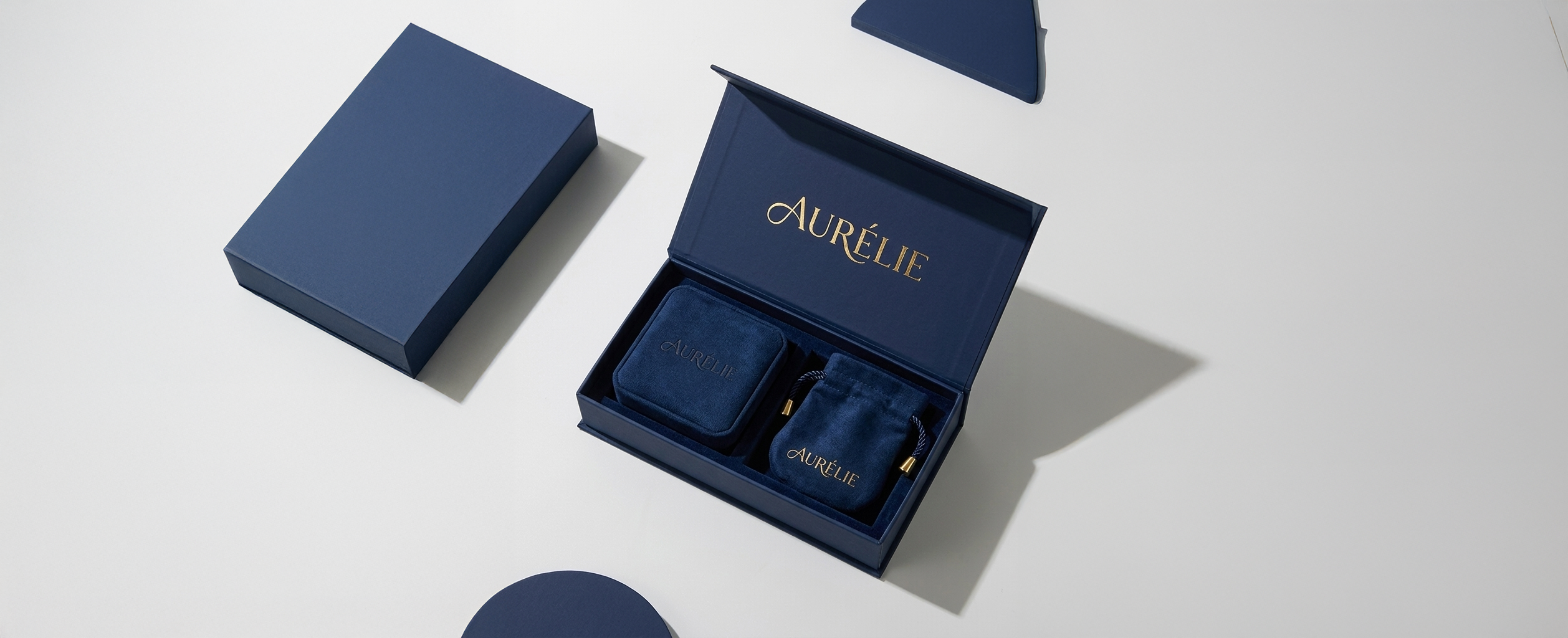 professional studio photograph showcases a premium, navy blue magnetic jewelry packaging suite manufactured by creativespacegifts. The centerpiece is an open rectangular rigid box featuring the brand name "AURÉLIE" elegantly stamped in gold foil on the inner lid. Inside, the box is meticulously organized with a matching navy velvet ring box and a soft drawstring pouch, both displaying the same gold-toned branding. To the left, a closed version of the box highlights its clean, matte finish and sturdy construction. The entire set is arranged on a minimalist white surface, accented by deep blue geometric shapes and soft directional shadows that emphasize the high-end, tactile quality of the materials.